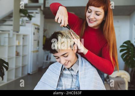 Cheerful female hairdresser with scissors making haircut to Woman with dyed hair in light room with green plants and stairway Stock Photo