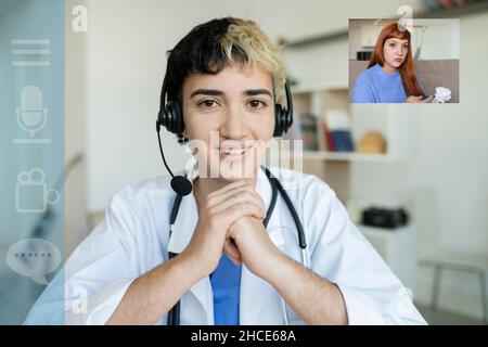Professional female medical operator with headset in uniform looking at camera while making video call to patient during online consultation Stock Photo