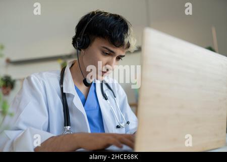 Focused female medical operator with headset in uniform and stethoscope browsing modern netbook at table during work in call center Stock Photo