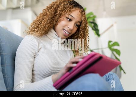 African American Woman sitting on sofa with crossed legs while resting in modern apartment with tablet Stock Photo