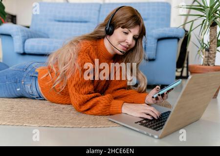 Content female operator in headset typing on netbook while lying on floor with smartphone in living room near couch during online work Stock Photo