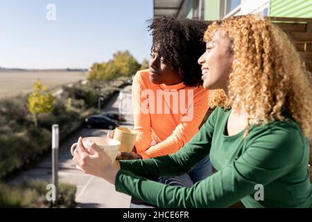 Side view of young multiracial female friends in casual clothes standing on balcony with coffee and talking Stock Photo