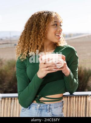 Young curly haired ethnic Woman resting on balcony with cup of coffee in daytime looking away Stock Photo