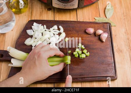 From above of crop anonymous person slicing fresh green celery with knife on wooden cutting board garlic and bay leaves placed on table for cooking co Stock Photo