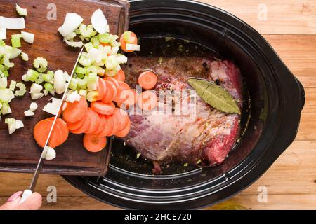 From above of crop anonymous person putting sliced carrot onion and celery from chopping board into slow cooker with beef with seasonings while prepar Stock Photo