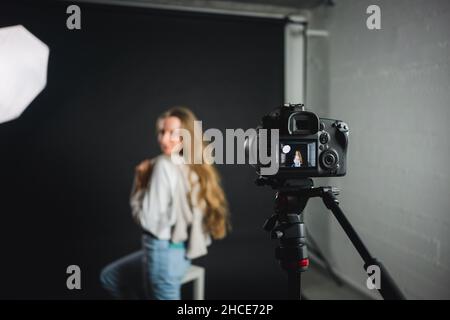 Side view of female with long hair in casual clothes standing in front of professional photo camera on tripod in studio Stock Photo