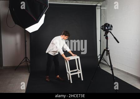 Full body of guy in casual clothes carrying white wooden chair on black background in professional photo studio Stock Photo