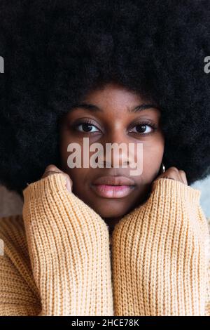 Portrait of cheerful young African American female with Afro hairstyle on a gray background with his hands hidden in his sweater resting on his face w Stock Photo