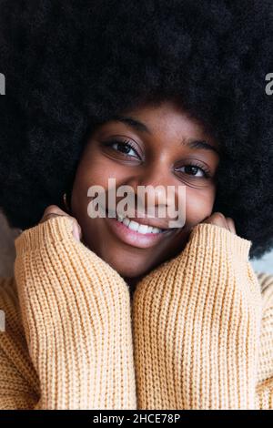 Portrait of cheerful young African American female with Afro hairstyle on a gray background with his hands hidden in his sweater resting on his face w Stock Photo