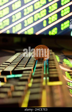 Round metal bitcoin coin placed on black keyboard of computer against blurred glowing monitor with numbers Stock Photo