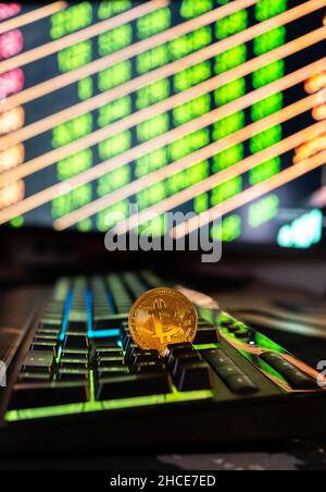Round metal bitcoin coin placed on black keyboard of computer against blurred glowing monitor with numbers Stock Photo