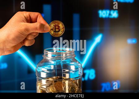 Crop anonymous man demonstrating bitcoin cryptocurrency holding it over glass jar with money against glowing financial chart background Stock Photo