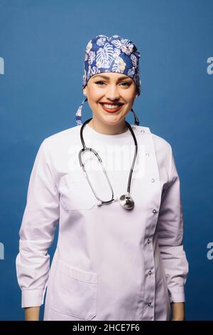 Positive doctor Woman in white uniform robe and bandana with stethoscope looking at camera on blue background Stock Photo