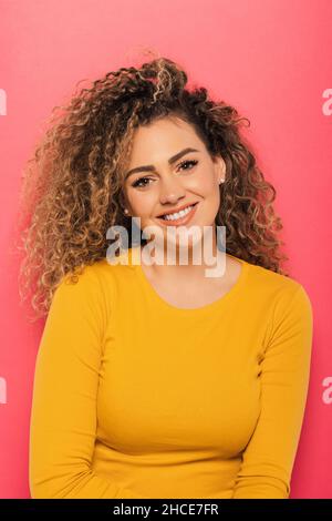 Young curly haired Woman in bright yellow sweater looking at camera on pink background Stock Photo