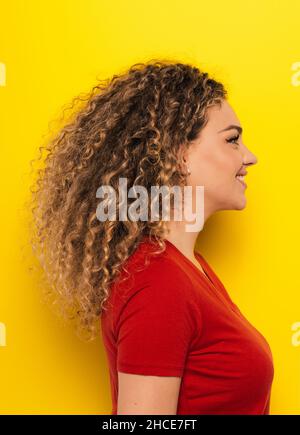Side view portrait of young curly haired Woman in bright red t-shirt looking away on yellow background Stock Photo