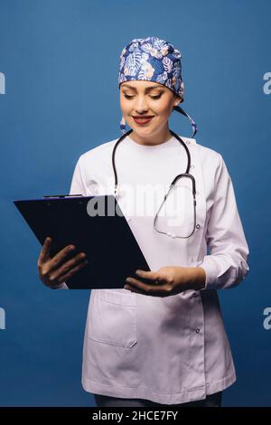 Positive doctor Woman in white uniform robe and bandana with stethoscope looking at clipboard tablet folder on blue background Stock Photo