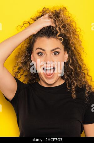 Young Woman in black t-shirt with amazed expression on yellow background grabbing her hair Stock Photo