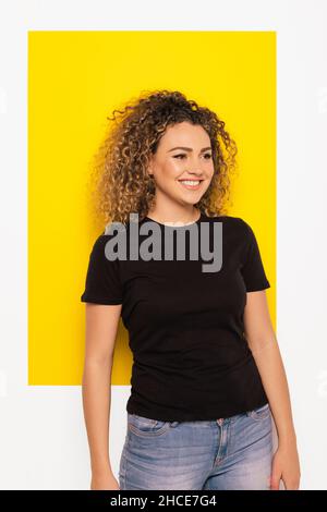 Young curly haired Woman in black t-shirt looking away on yellow and white background Stock Photo