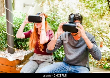 Young couple sitting on wooden bench and experiencing another virtual world in headset in park against green plants Stock Photo