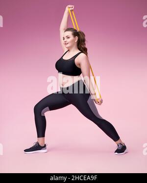 Strong girl working with resistance band. Photo of model with curvy figure in fashionable sportswear on pink background. Sports motivation and healthy Stock Photo