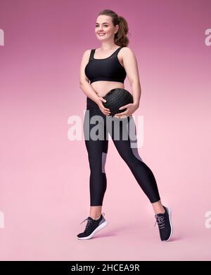 Sporty girl with medicine ball. Photo of model with curvy figure in fashionable sportswear on pink background. Dynamic movement. Sports motivation and