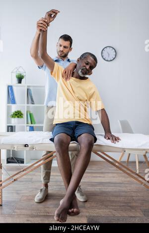 physiotherapist raising hand of african american patient sitting on massage table Stock Photo