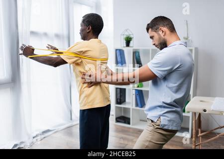 young trainer supporting mature african american man working out with elastics Stock Photo