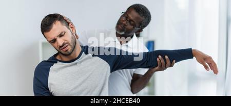 young man frowning from pain while african american rehabilitologist raising his arm, banner Stock Photo