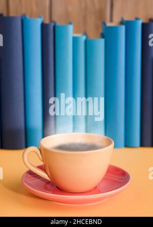 Yellow cup of coffee or tea with hot steam, on book with light blue books and yellow table background. Stock Photo