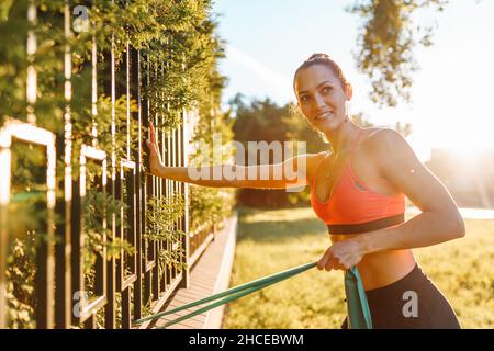 Young beautiful sporty woman in black and pink top and black shorts exercising using resistance rubber band on fence and smiling at sunrise in summer Stock Photo