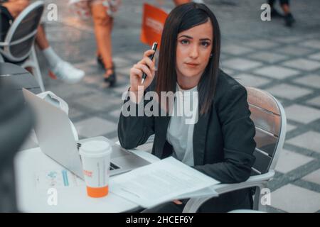 Young lady secretary making notes with her laptop and reports on sheets of paper at the table along with a coffee in the cafeteria in the city center Stock Photo