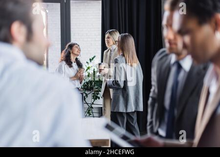 smiling businesswomen with coffee to go talking near blurred multiethnic businessmen in office Stock Photo
