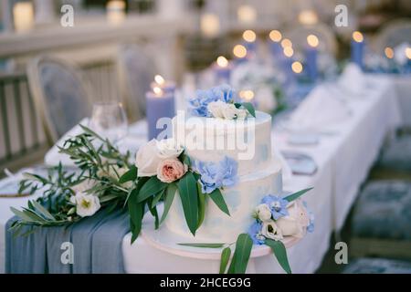 White bunk wedding cake decorated with flowers and leaves stands on a table with candles Stock Photo
