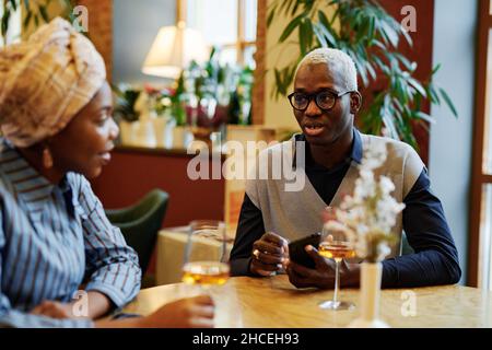 African young man in eyeglasses sitting at the table and talking to woman they drinking wine during meeting in cafe Stock Photo