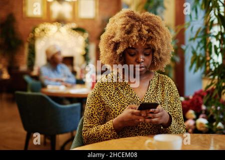 African young woman with elegant hairstyle using mobile phone while sitting at the table in cafe Stock Photo