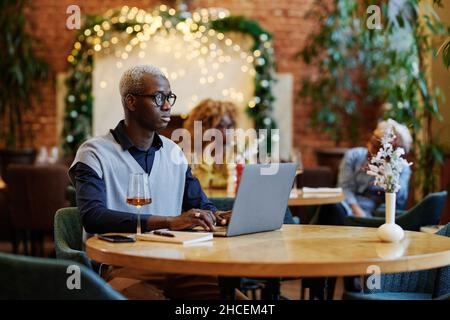 Serious young businessman in eyeglasses working on laptop at the table with glass of white wine, he working in cafe Stock Photo