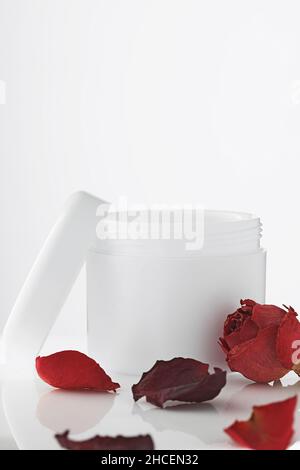 Empty white plastic jar with lid open on light background. Petals of red roses. Rejuvenating cosmetics. Care product for skin of body and face. Layout for logo. Cosmetology and spa. Stock Photo