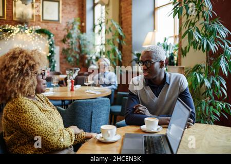 African young man talking to his colleague while they drinking coffee at the table during their business meeting in cafe Stock Photo