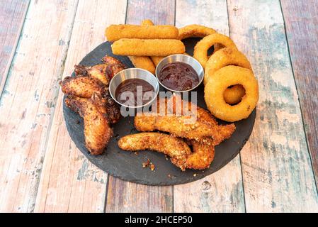 Tex Mex appetizers with onion rings, cheese sticks, chicken wings, BBQ wings and sauces on a round black slate plate Stock Photo