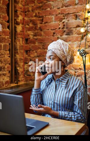 African serious woman having a conversation on mobile phone while working on laptop at the table in cafe Stock Photo