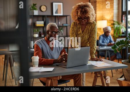 African young businesswoman looking at computer monitor and talking to businessman while he typing on laptop at the table Stock Photo