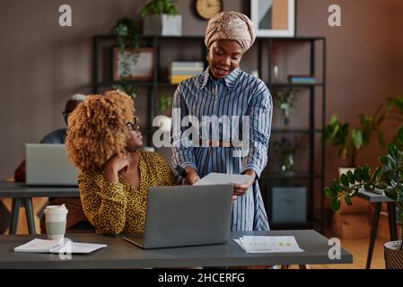 African businesswoman reading a document and discussing it with her colleague while she working on laptop, they working in team at office Stock Photo