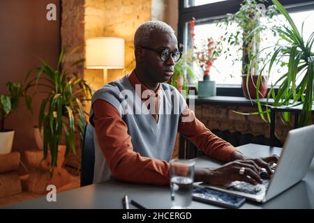 Serious African businessman in eyeglasses sitting at the table and typing on laptop working at office Stock Photo