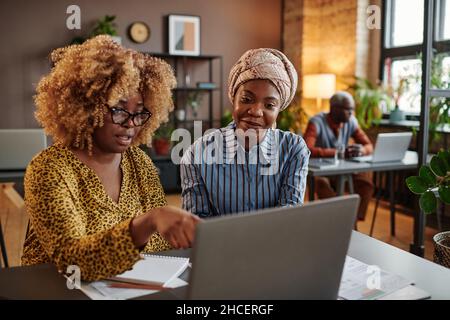African businesswoman with curly hair pointing at laptop while sitting at the table with her colleague and they working in team at office Stock Photo