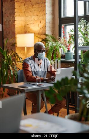 African young businessman in eyeglasses wearing protective mask working at his workplace on laptop at office Stock Photo
