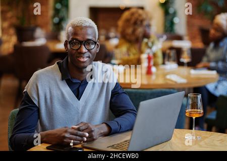 Portrait of African young businessman in eyeglasses smiling at camera while sitting at the table with laptop and glass of wine Stock Photo