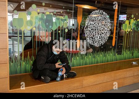 Young man sitting crosslegged on a windowsill in the departures lounge looking at his smart phone and wearing a facemask, Dublin Airport, Dublin, Irel Stock Photo