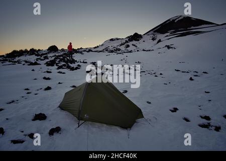 man and his tent at dawn and moonset camping on the snow by a crater of Etna Mountain in Sicily Stock Photo