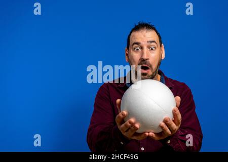 Spanish bearded man holding a white ball totally amazed and excited to behold it, isolated on blue studio background Stock Photo
