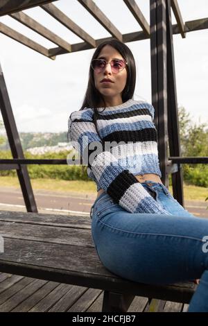 beautiful young latin model posing with black straight hair in a park with nature, wears sunglasses, jeans and casual sweater, beauty and fashion Stock Photo